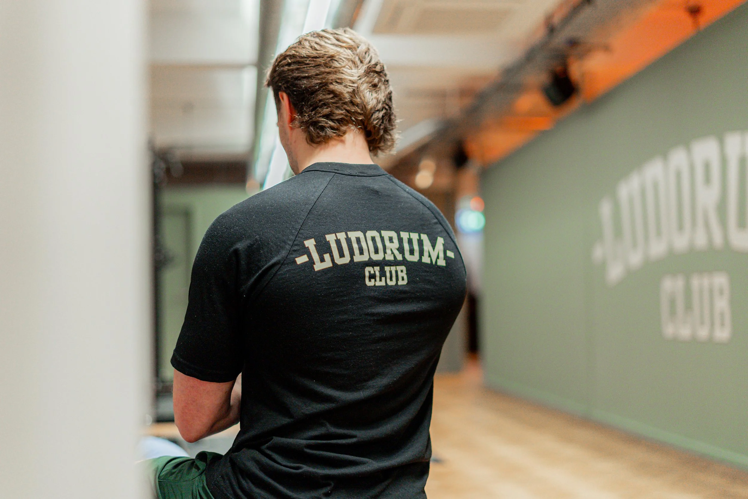 Jake from behind in a black Ludorum Club T shirt, the painted wall out of focus beyond